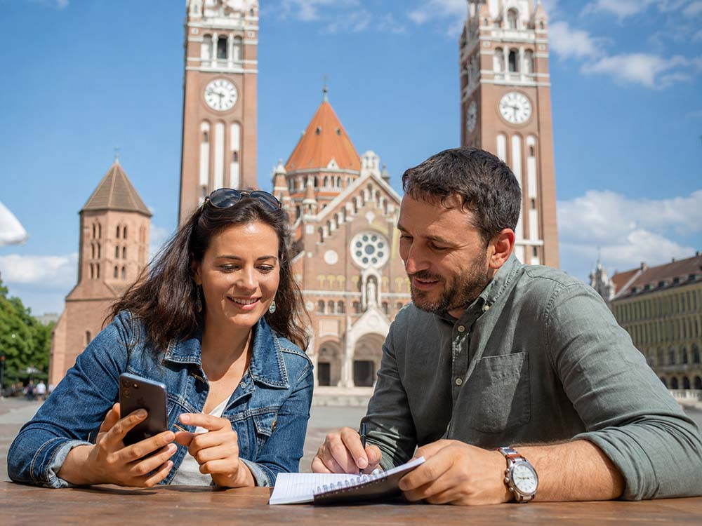 Paar lernt Ungarisch mit Smartphone und Notizbuch vor dem Dom in Szeged